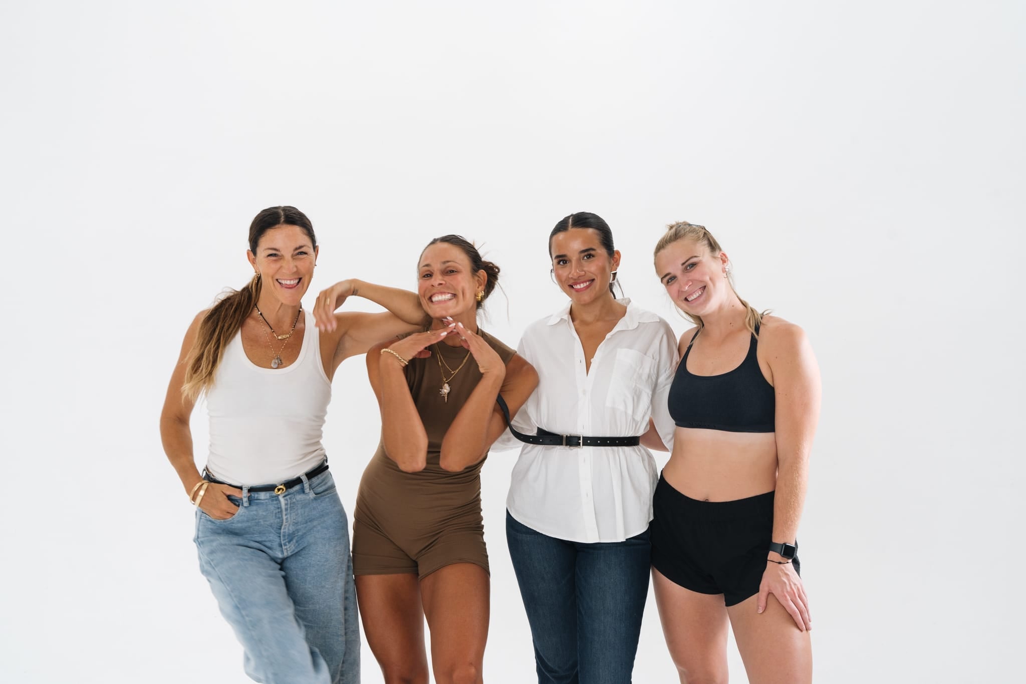 Four women standing together against a plain white background, smiling and posing casually; two wear casual outfits, two wear activewear.